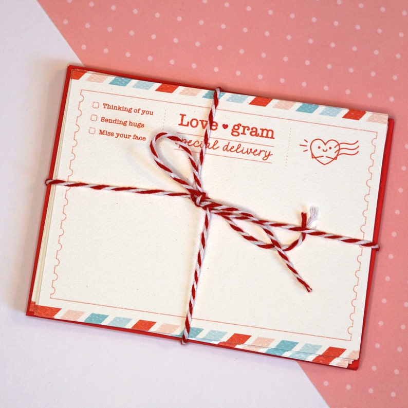 May include: A set of blank cards with a red, white, and blue border. The cards are tied together with a red and white twine. The cards have the text "Love gram special delivery" and a cartoon heart with wings. The cards have checkboxes with the text "Thinking of you", "Sending hugs", and "Miss your face".