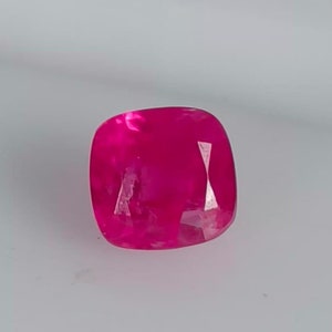 May include: A vibrant pink gemstone with a cushion cut. The gemstone has a smooth, polished surface and is set against a white background.
