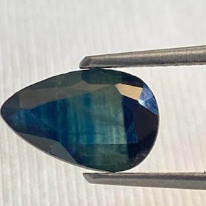 Può includere: Una pietra preziosa a forma di pera con un colore blu-verde profondo. La pietra preziosa è tenuta in una pinzetta su uno sfondo bianco.