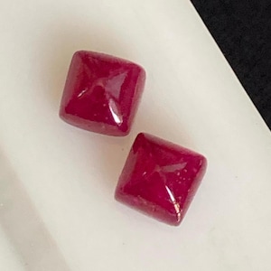May include: Two square-cut, deep red gemstones with rounded corners. The gems have a polished, reflective surface, and are set against a white and black background. The gemstones are likely rubies, suitable for jewellery making.