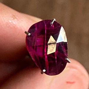 Könnte beinhalten: Ein facettierter, tropfenförmiger Edelstein in tiefem Magenta. Der Stein wird von drei silbernen Zinken gehalten. Der Schliff des Steins erzeugt mehrere reflektierende Oberflächen. Der Hintergrund ist ein verschwommenes, warmes Braun.
