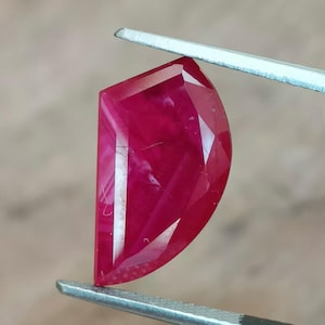 May include: A faceted, half-moon shaped ruby gemstone, held by tweezers. The ruby is a deep, saturated red colour with internal striations. The gem's cut reveals multiple facets, reflecting light. The tweezers are silver and black.