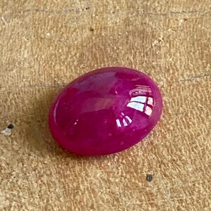 May include: A polished, oval-shaped, deep magenta gemstone. The gem reflects light, showcasing its smooth, glossy surface. The stone is set against a textured, light brown wooden surface.