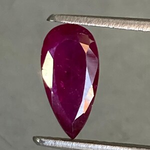 Può includere: Una pietra preziosa di forma a pera di colore rosso intenso tenuta da una pinzetta.
