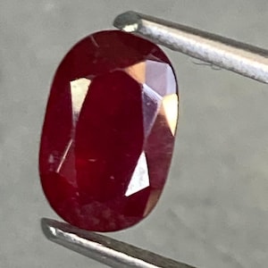 Op de afbeelding: Een dieprode ovale edelsteen vastgehouden door een pincet. De edelsteen is gefacetteerd en heeft een hoge glans.