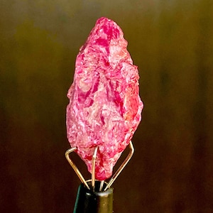 May include: A vibrant pink ruby crystal, with a rough, faceted surface, is held in a metal clamp.