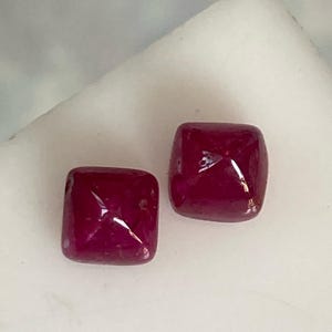 Könnte beinhalten: Zwei quadratische, tiefrote Edelsteine. Die Edelsteine sind facettiert und haben eine glänzende Oberfläche.