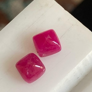 May include: Two square-shaped pink gemstones, likely rubies, are displayed on a white surface. The gemstones are smooth and polished, with a slight shine.