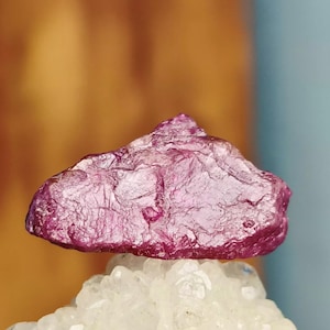 May include: A deep pink, rough-cut ruby crystal with a natural, irregular shape. The crystal is sitting on a bed of white, sparkly crystals.
