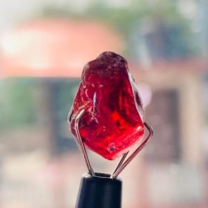 Könnte beinhalten: Ein roher, tiefroter Edelstein, der von einem Metall- und schwarzen Werkzeug gehalten wird. Der Edelstein hat eine raue, strukturierte Oberfläche und eine durchscheinende Qualität, die das Licht einfängt. Der Hintergrund ist verschwommen und deutet auf eine Umgebung im Freien hin.