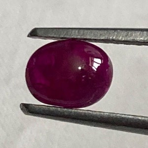 May include: A deep red ruby gemstone, held by a pair of tweezers, with a smooth, polished surface.