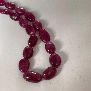 Puede incluir: Un hilo de cuentas de rubí de color magenta intenso, con forma ovalada y dispuestas en una línea curva. Cada cuenta presenta una superficie lisa y brillante con variaciones naturales de color y textura, reflejando la luz en sus formas.