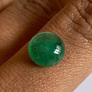 Könnte beinhalten: Ein runder, polierter Smaragd-Edelstein. Der Edelstein hat eine tiefe, lebendige grüne Farbe und eine glatte, reflektierende Oberfläche. Der Smaragd wird in einer Hand gehalten, um die Größe und Farbe des Edelsteins zu zeigen.