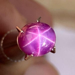 Puede incluir: Una piedra preciosa de zafiro estrellado rosa con una estrella de seis puntas en el centro.