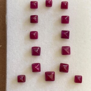 Puede incluir: Una colección de 13 piedras preciosas cuadradas de color rojo rubí con una parte superior en forma de pirámide. Las piedras preciosas están dispuestas en dos filas sobre un fondo blanco.