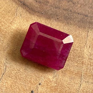 Op de afbeelding: Een gefacetteerde, rechthoekige robijn edelsteen met een dieprode kleur. De edelsteen heeft een gepolijst oppervlak en scherpe randen, die het licht reflecteren. De robijn ligt op een getextureerd, lichtbruin houten oppervlak.
