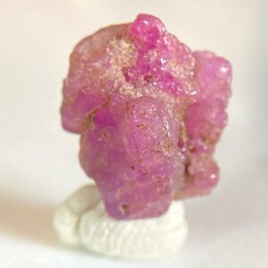 May include: A raw, unpolished ruby gemstone, exhibiting a vivid pink colour. The natural crystal structure is set on a small, white, textured base. The gemstone's surface has a slightly granular texture.