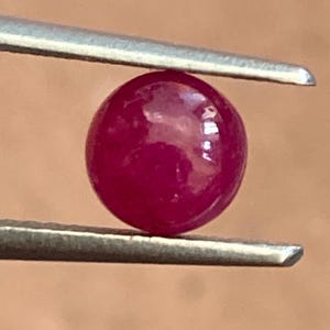 May include: A round, polished ruby gemstone held by tweezers. The ruby is a deep, saturated red color with a smooth, reflective surface. The tweezers are silver-colored and provide a contrasting backdrop to the vibrant gemstone.