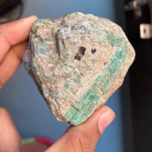 May include: A rough, green emerald crystal with a light green and white matrix. The crystal has a dark green color and is surrounded by a lighter green and white matrix.