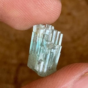 以下が含まれることがあります： 柱状構造の小さな水色の結晶クラスター。 結晶は半透明で、内部の縞模様とわずかに粗い質感が見えます。 結晶は2本の指で持たれており、そのサイズと形状を示しています。