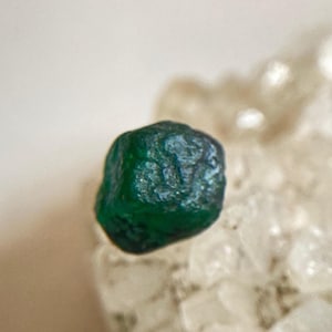 Pode incluir: Um close-up de uma gema de esmeralda bruta, verde escuro. A esmeralda tem uma superfície texturizada e é colocada sobre um fundo de um material cristalino mais claro. A gema é o ponto focal.