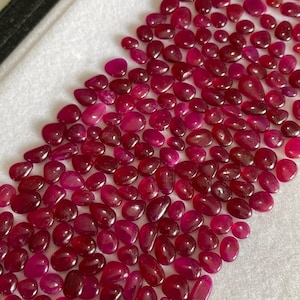 May include: A collection of polished, oval-shaped ruby gemstones in varying sizes and shades of red. The gemstones are scattered on a white surface, with a few darker red stones mixed in.