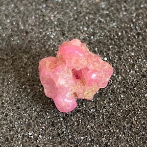 Könnte beinhalten: Ein roher, rosa Edelstein mit kristalliner Struktur. Der Stein hat eine unregelmäßige Form und einige kleine Hohlräume. Die Farbe ist ein leuchtendes, helles Rosa mit einigen Bereichen von etwas dunklerem Farbton. Der Edelstein liegt auf einem dunklen, strukturierten Hintergrund.
