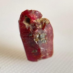 Pode incluir: Uma gema de rubi vermelho escuro em bruto. A pedra natural tem uma forma ligeiramente irregular com uma superfície texturizada e inclusões visíveis. A cor do rubi é um vermelho rico e saturado.