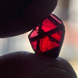 Puede incluir: Una piedra preciosa de color rojo intenso y translúcido con una forma geométrica facetada. La piedra muestra inclusiones oscuras internas y fracturas, captando la luz para revelar un tono carmesí vibrante en su estructura.
