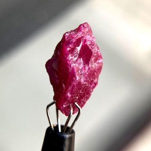 Könnte beinhalten: Ein roher, tief magentafarbener Edelstein mit einer gezackten, kristallinen Textur. Der Stein wird von einem schwarzen Metallwerkzeug mit vier dünnen, silberfarbenen Drahtarmen aufrecht gehalten, die die Basis des unregelmäßigen Minerals greifen.