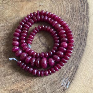 Op de afbeelding: Spiraalvormige dieprode robijn kralen. Elke kraal is afgerond en geregen, waardoor een ketting of armband ontstaat. De achtergrond is een lichtbruin houten oppervlak. De kralen zijn ongeveer 0,5 cm in diameter.