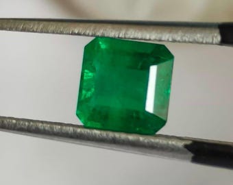 Puede incluir: Una gema de esmeralda verde vibrante, cortada en forma cuadrada con bordes biselados. La esmeralda está sujeta por unas pinzas.