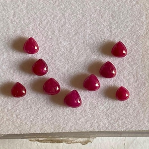 May include: Eight small, loose, teardrop-shaped rubies with a deep red colour and a smooth, polished finish. The gemstones are arranged in a scattered, semi-circular pattern, showcasing their vibrant hue and consistent pear-cut shape.