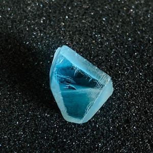 Puede incluir: Una gema facetada de color azul claro con una estructura cristalina. La gema tiene una cualidad translúcida, permitiendo el paso de la luz. La piedra se encuentra sobre un fondo oscuro y texturizado, resaltando su color y forma. La gema parece ser un mineral natural.