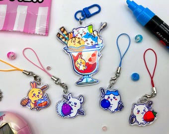 May include: A collection of colorful acrylic charms featuring cute cartoon characters and food-themed designs. The charms include a sundae, bunnies, a mouse with berries, and a cat with a blueberry. Each charm has a lanyard for attachment.