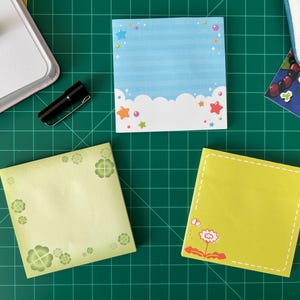 May include: An assortment of colorful sticky notes with various designs, including a blue sky with clouds, four-leaf clovers, and a flower. Other items include a small black flashlight, a green beaded bracelet, and star-shaped green earrings.