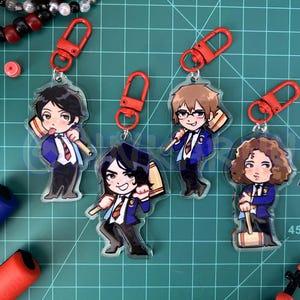 May include: Five acrylic anime-style character keychains with red clasps. Each keychain features a different character in a blue blazer, white shirt, and red tie, holding a wooden mallet. The background is a green cutting mat.