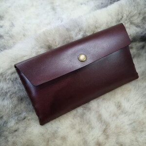 Op de afbeelding: Een bordeauxrode leren clutch met een klep en een goudkleurige knoop. De rechthoekige portemonnee is gemaakt van glad leer en wordt getoond op een zacht, lichtgekleurd oppervlak.