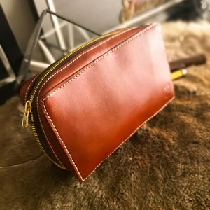 May include: A brown leather zippered pouch with white stitching. The pouch is lying on a brown fuzzy surface.