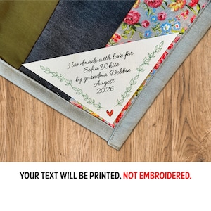 May include: A personalised label with the text "Handmade with love for Sofia White by grandma Debbie August 2026" is sewn onto a patchwork quilt. The label is surrounded by a green leaf design and a small red heart. The quilt has various fabric patterns.