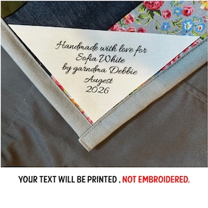 May include: A white triangular label with black script reading "Handmade with love for Sofia White by garndma Debbie August 2026." The label is sewn onto a patchwork quilt with floral and denim fabrics. The text "YOUR TEXT WILL BE PRINTED, NOT EMBROIDERED" is at the bottom.