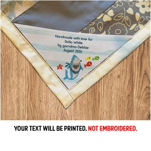 May include: A handmade baby blanket with a shark and fish design. The blanket features a label that reads "Handmade with love for Sofia White by grandma Debbie August 2026." The text "YOUR TEXT WILL BE PRINTED, NOT EMBROIDERED" is also visible.
