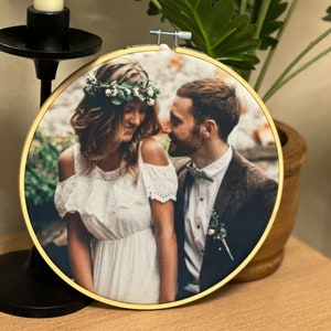 Puede incluir: Un aro de bordado redondo con una imagen impresa de una novia y un novio. La novia lleva un vestido blanco con una corona de flores y el novio lleva un traje y una corbata de lazo. La imagen está sobre un fondo de vegetación.