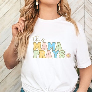 Może przedstawiać: Biała koszulka z napisem "This Mama Prays" w kolorowych literach blokowych z małym wzorem kwiatka.