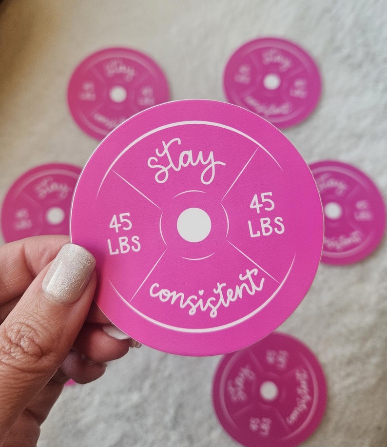 Op de afbeelding: Ronde roze sticker met een wit gewichtsschijf ontwerp. De sticker zegt "Stay consistent" en "45 LBS" aan elke kant van de schijf.