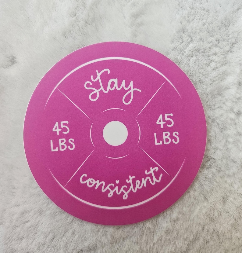 Op de afbeelding: Ronde roze gewichtsplaat met witte tekst die "Stay consistent" en "45 LBS" aan beide kanten van de plaat zegt.