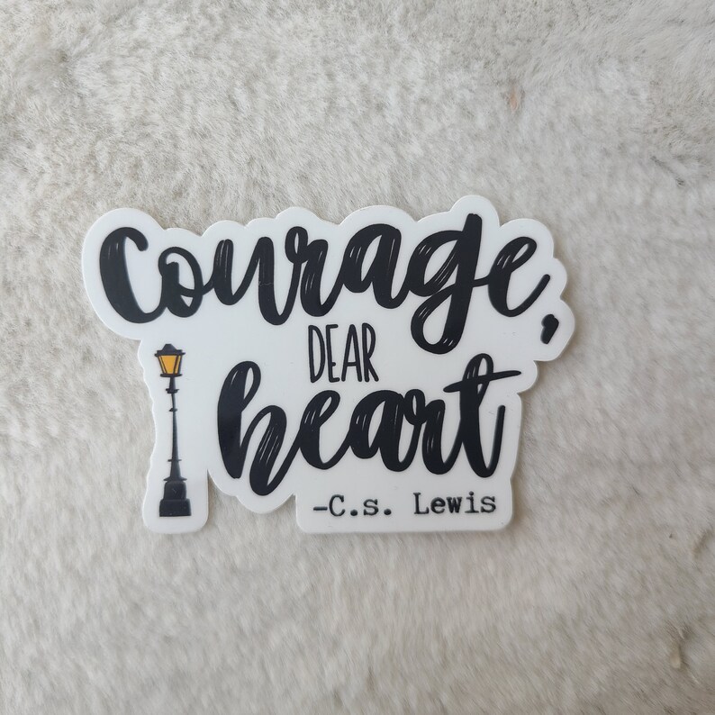 May include: Black and white sticker with the quote "Courage, dear heart - C.S. Lewis" in a handwritten font. A small black silhouette of a street lamp with a yellow light is to the left of the text.