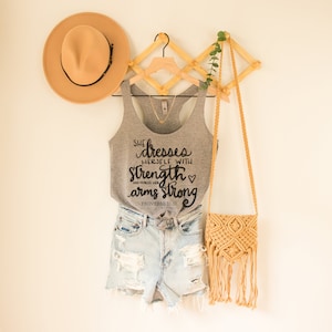 May include: Gray tank top with the text "She dresses herself with strength and makes her arms strong - Proverbs 31:17" in black lettering. The tank top is hanging on a wooden hanger with a straw hat and a woven bag.