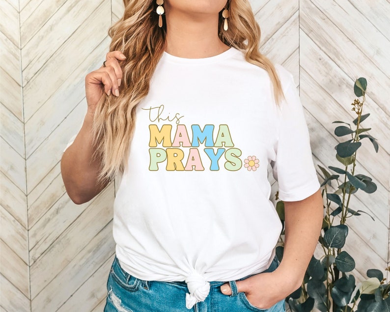 Może przedstawiać: Biała koszulka z napisem "This Mama Prays" w kolorowych literach blokowych z małym wzorem kwiatka.