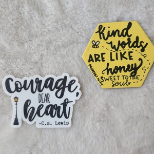 May include: Two yellow and black motivational stickers. The first sticker is a hexagon with the text "Kind words are like honey sweet to the soul" and a small bee illustration. The second sticker is a white background with black text that reads "Courage, dear heart - C.S. Lewis" and a black silhouette of a street lamp.